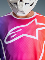 Alpinestars 2026 Fluid Apex tröja, motocrosströja, mångfärgad lila, rosa och orange gradient, med en stor vit Astars-logotyp på bröstet, närbild bärs tillsammans med en vit motocrosshjälm