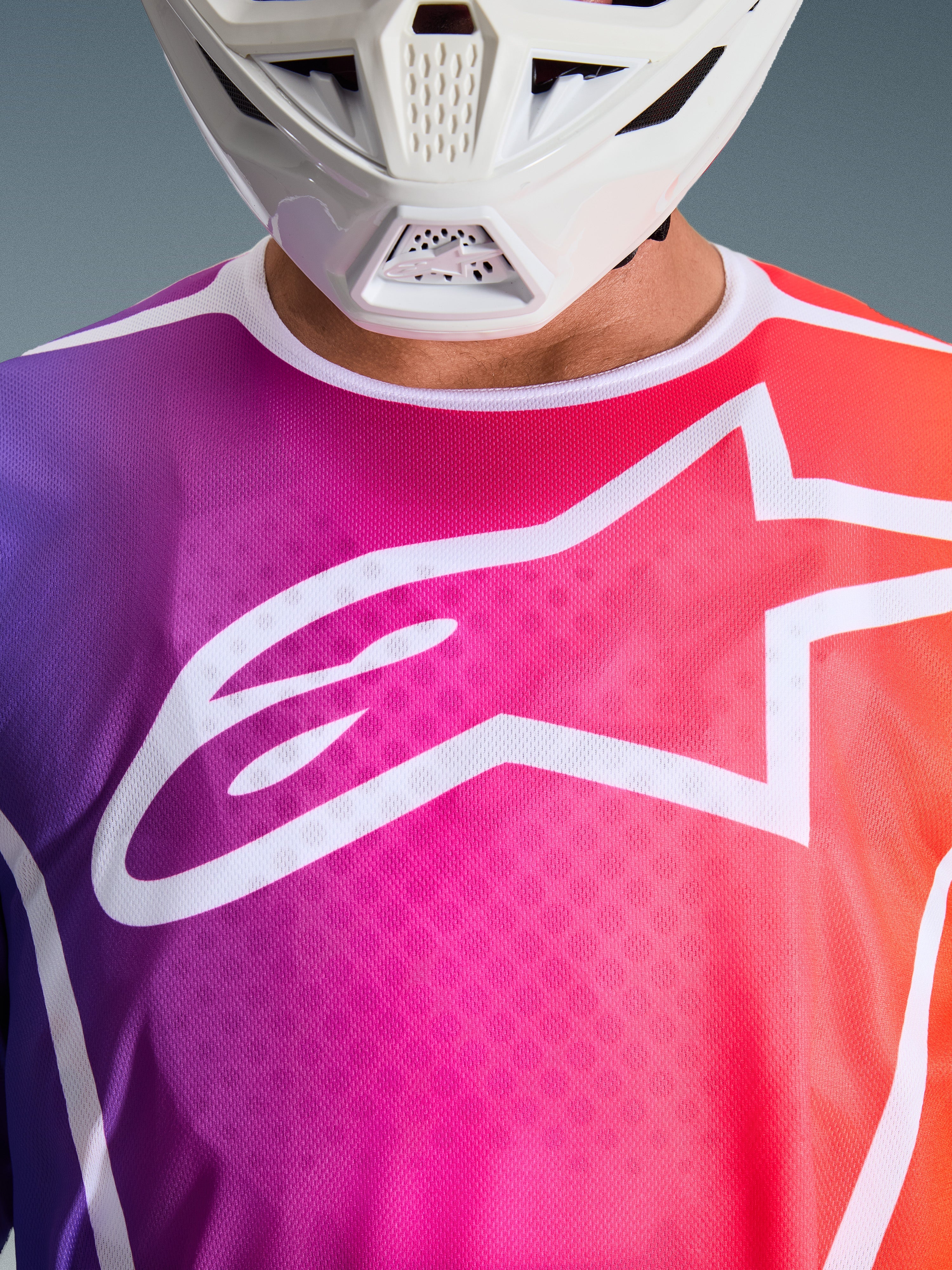Alpinestars 2026 Fluid Apex Jersey, motocrosströja, färgtoning i lila, rosa och orange, stor vit Alpinestars-stjärnlogotyp på bröstet, närbild som visar vitt hakskydd från hjälm