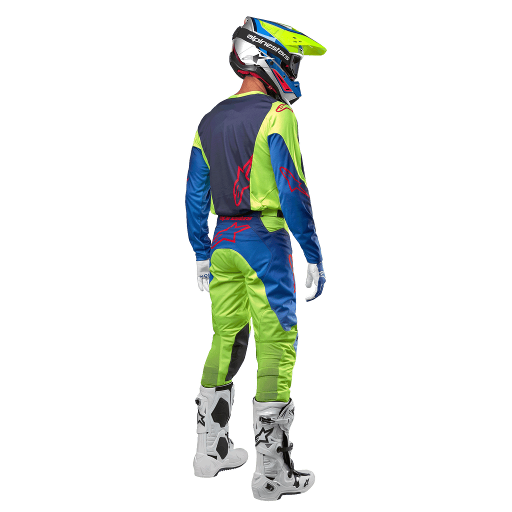 Motocrossförare sedd bakifrån i Alpinestars 2024 Racer Hoen-tröja i Yellow Fluo och Blue Night Navy, matchande motocrossbyxor och vita stövlar, visar ryggdesignen med röda logotyper, koordinerad outfit för professionell terrängkörning