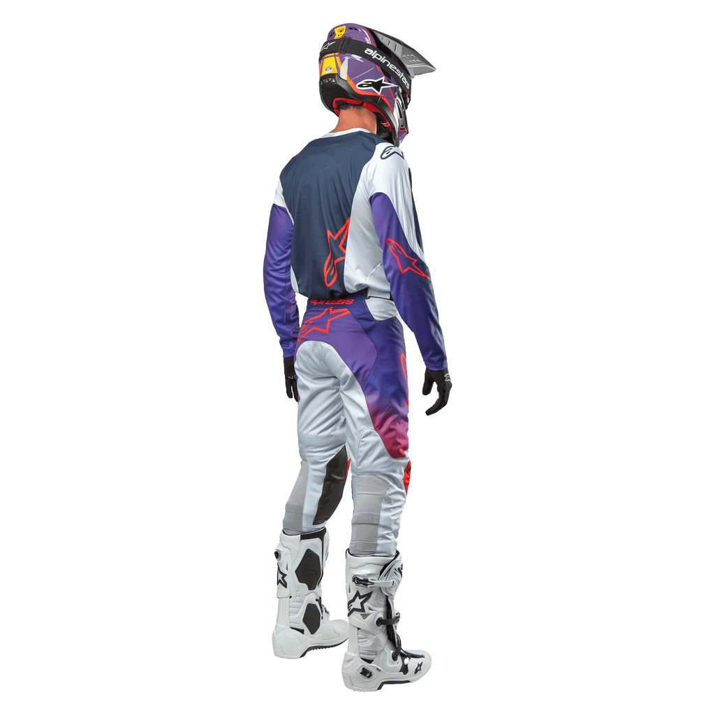 Motocrossförare sedd bakifrån bärande Alpinestars 2024 Racer Hoen Jersey i Light Gray Hot Orange Black, mörkblå ryggpanel med orange stjärnlogotyp, lila ärmar, bärs tillsammans med matchande racingbyxor och vita stövlar.