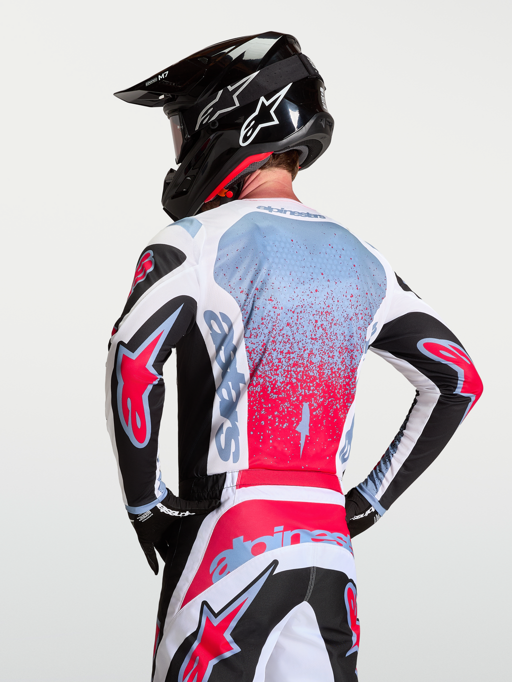 Alpinestars Fluid Drag tröja, motocrosströja i svart, vitt och rött med ljusblå detaljer, bärs av modell bakifrån med matchande byxor och hjälm, andningsbar polyfabrikskonstruktion för terrängkörning.
