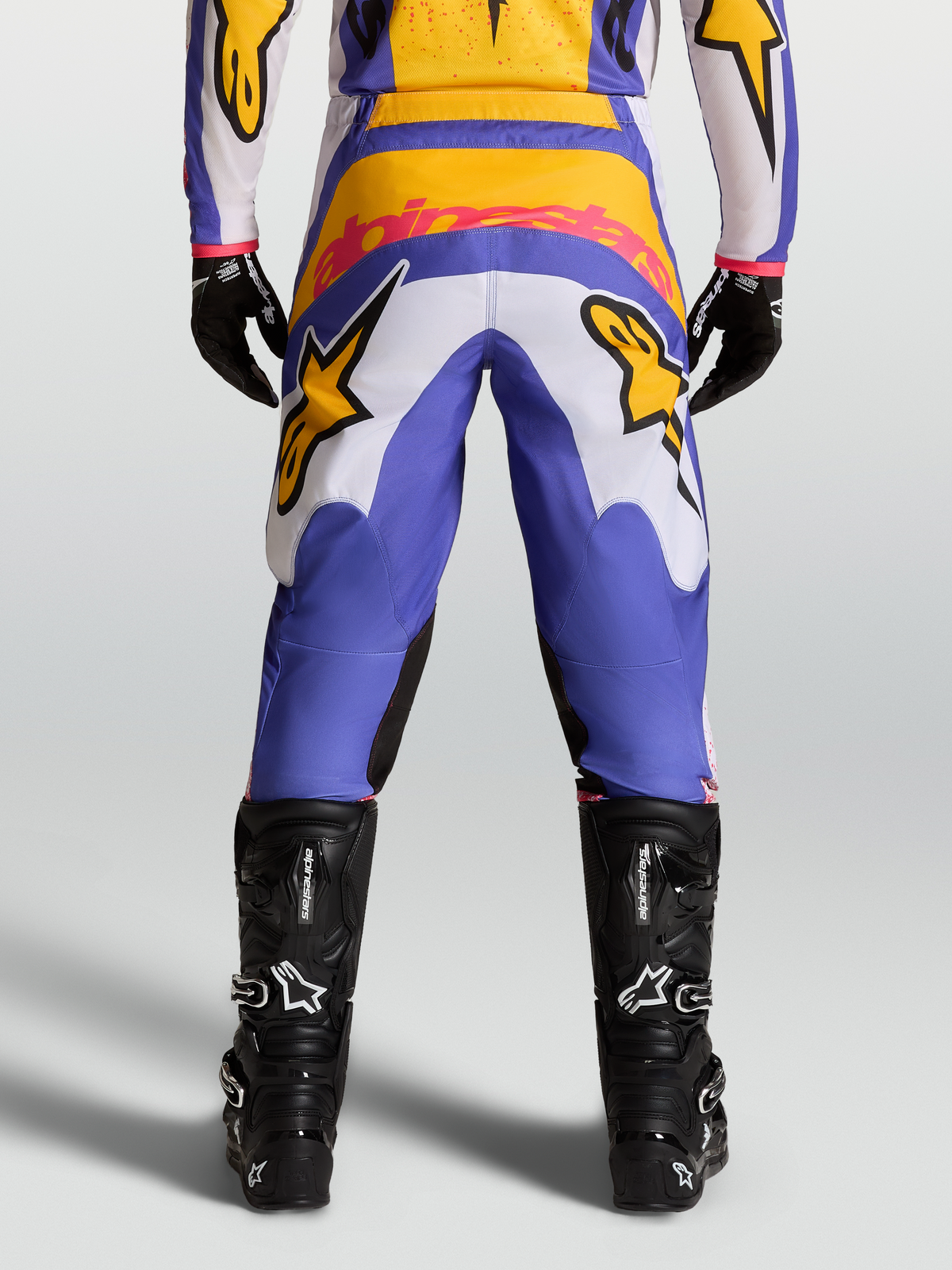 Alpinestars Fluid Drag Pants for motocross racing, view from behind, in Blue and Gold with stretch corduroy panels and reinforced seat, paired with black technical boots.