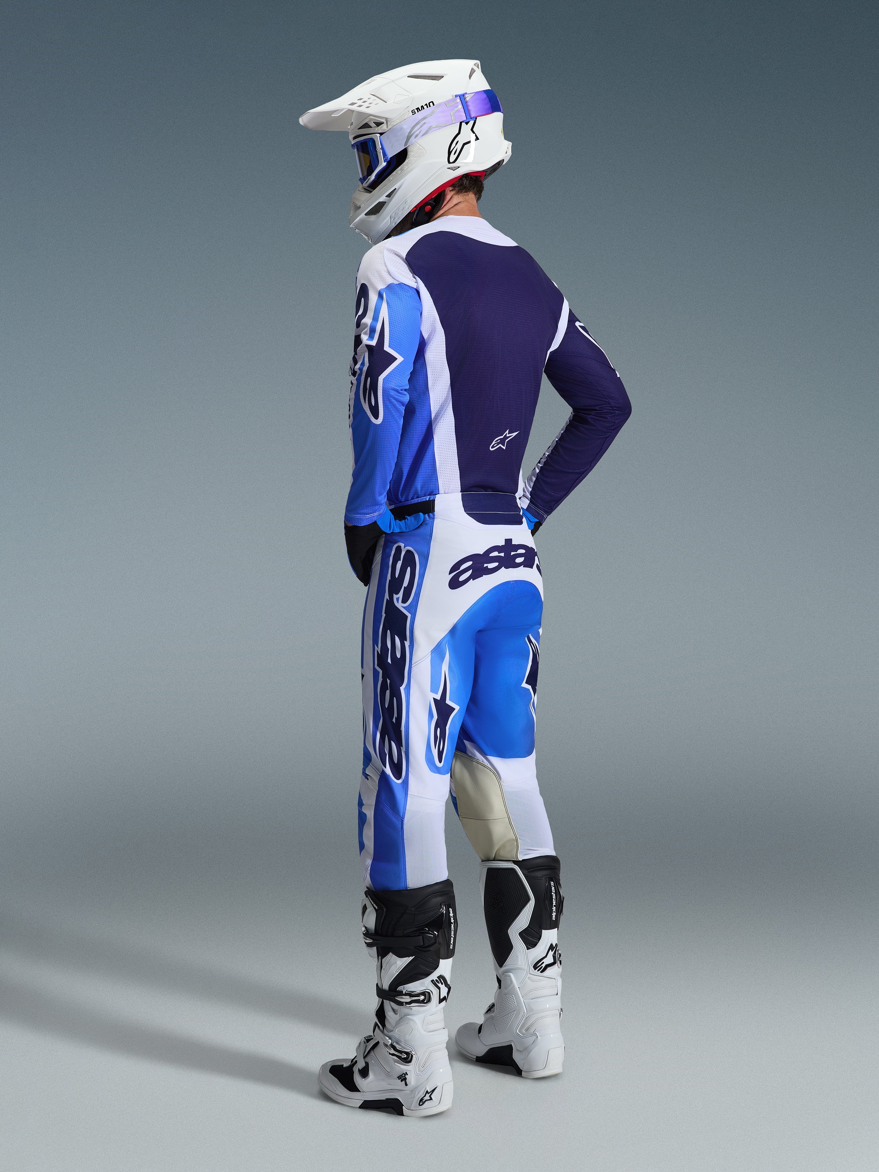 Motocross rider wearing the Alpinestars 2026 Racer Air Portl Gear Set from a rear view, blue and white jersey with dark navy accents, matching pants with Astars logos, white helmet with blue goggles, white and black boots, coordinated racing outfit for off-road performance