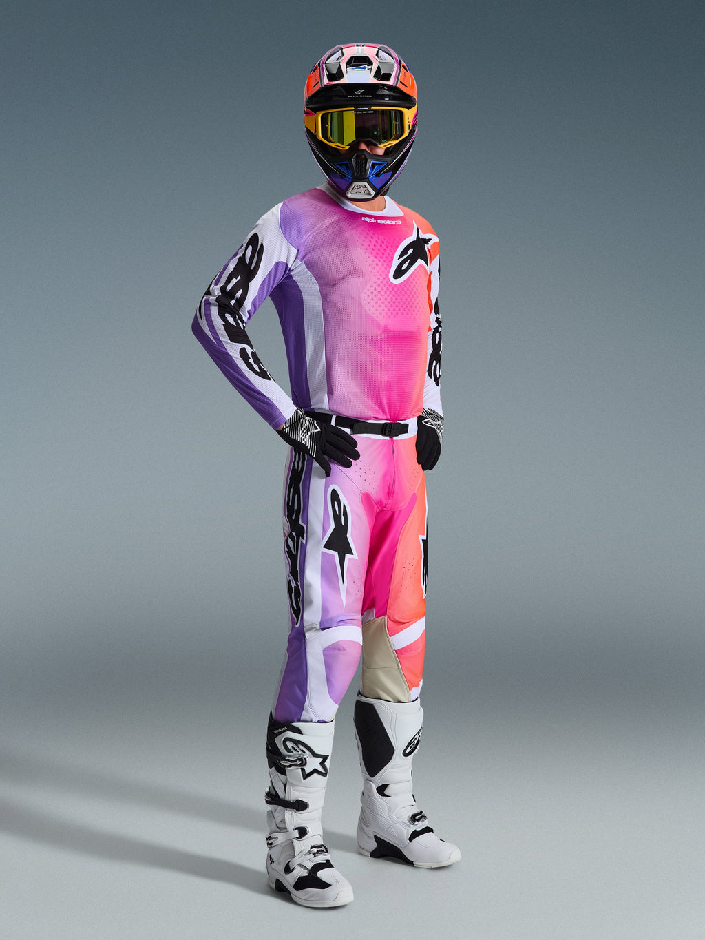 Alpinestars 2026 Racer Air Portl utrustningsset, motocrossförare i komplett racingutrustning, tröja och byxor med en livfull gradient i lila, rosa och orange, svarta Alpinestars-logotyper, vita och svarta detaljer, matchande flerfärgad hjälm med gultonade goggles, vita och svarta motocross-stövlar, svarta och vita handskar