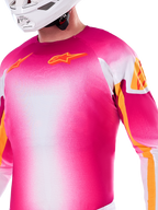 Alpinestars Limited Edition Supertech AFD MIG-tröja, motocross-tröja i en gradient av neonrosa och vitt, med orangea Astars-logotyper och detaljer på ärmarna, bärs av en förare med vit hjälm, atletisk racingpassform.