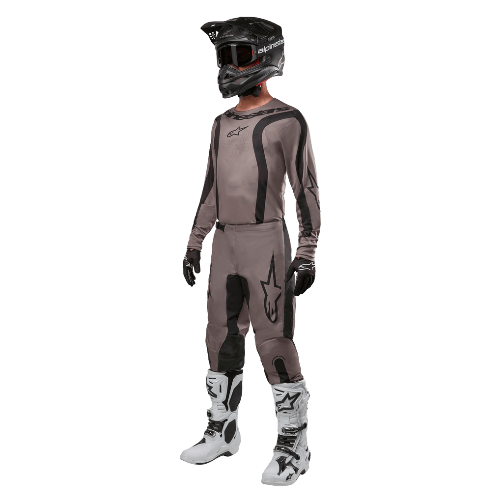 Motocrossförare i komplett racingutrustning med Alpinestars 2024 Fluid Lurv-tröja i Mud Black, matchande byxor, svart hjälm med goggles, vita och svarta stövlar och svarta handskar, koordinerad outfit för terrängkörning