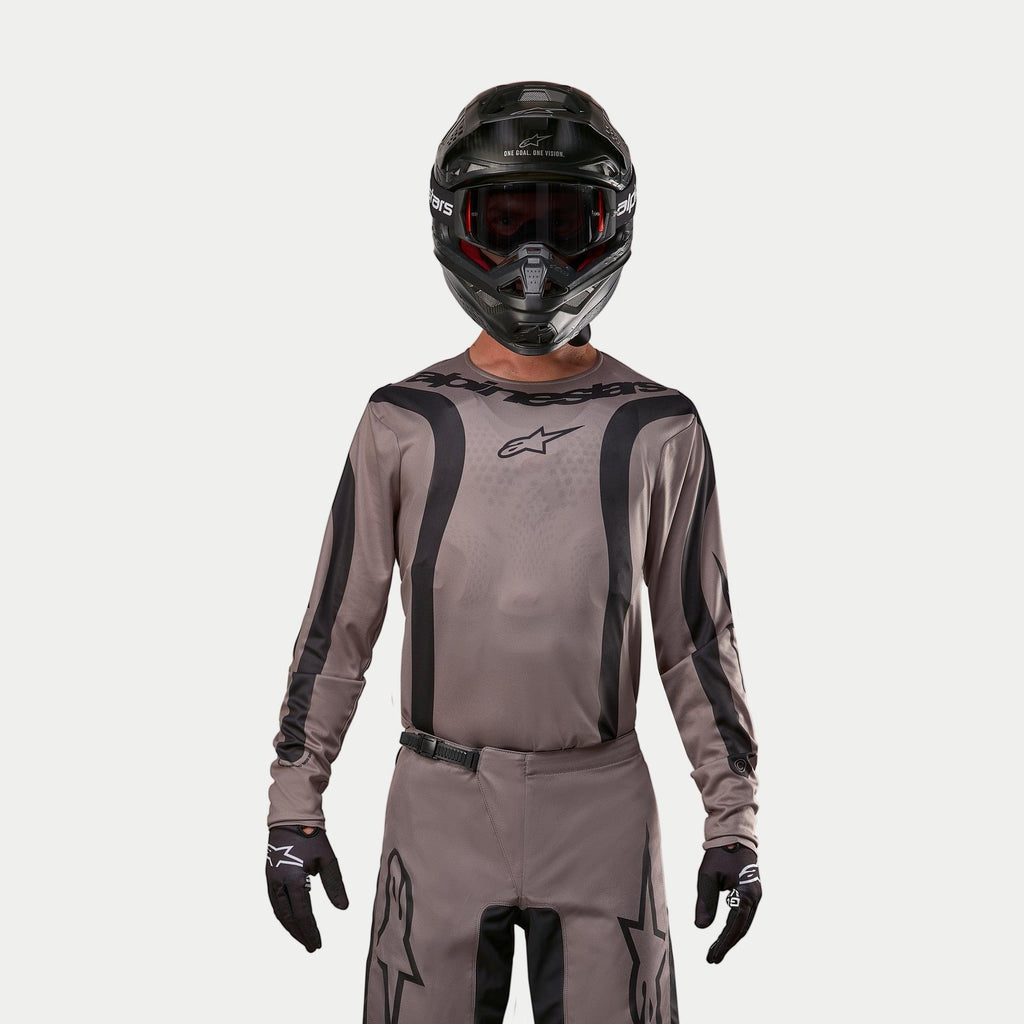 Motocrossförare iklädd Alpinestars 2024 Fluid Lurv-tröja i Mud Black, ett jordnära gråbrunt tyg med svarta grafiska ränder och en stjärnlogotyp på bröstet, kombinerat med en svart hjälm, röda goggles och svarta handskar för en koordinerad look för terrängkörning