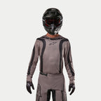 Motocrossförare iklädd Alpinestars 2024 Fluid Lurv-tröja i Mud Black, ett jordnära gråbrunt tyg med svarta grafiska ränder och en stjärnlogotyp på bröstet, kombinerat med en svart hjälm, röda goggles och svarta handskar för en koordinerad look för terrängkörning