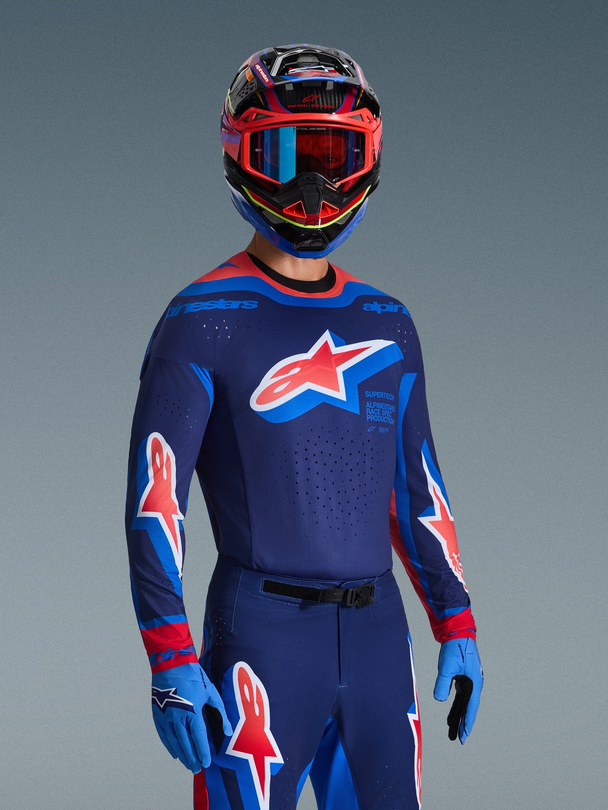Motocross rider wearing 2026 Supertech Vista Jersey in Dark Navy, Hot Coral, and Cobalt, matching pants and gloves, multi-colored helmet with red goggles, professional gear set for off-road racing
