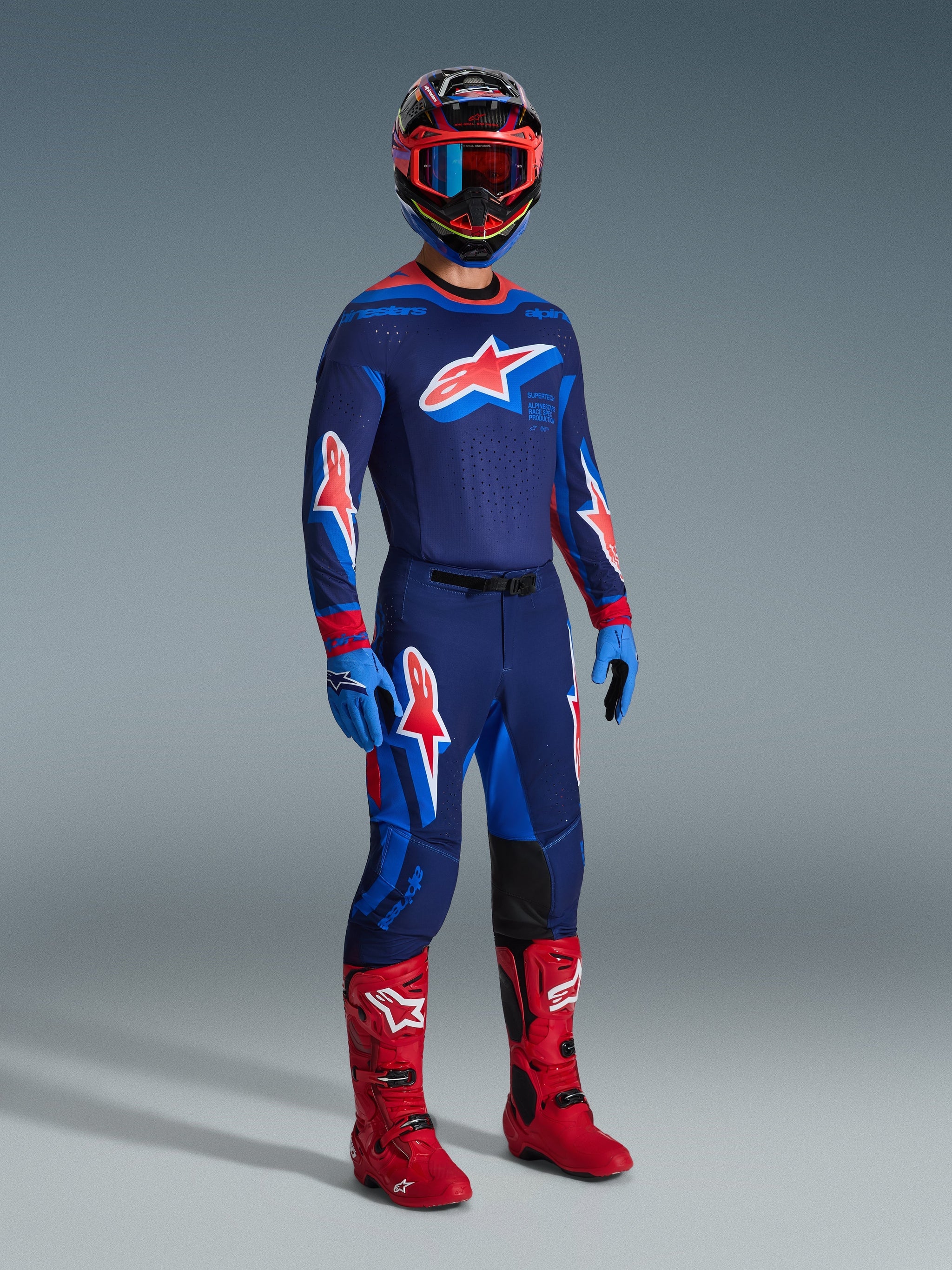Motocross rider wearing complete racing gear, featuring the Alpinestars 2026 Supertech Vista Jersey in Dark Navy, Hot Coral, and Cobalt, matching pants with bold star logos, bright red motocross boots, blue gloves, and a multi-colored helmet with red goggles, professional off-road racing gear set.
