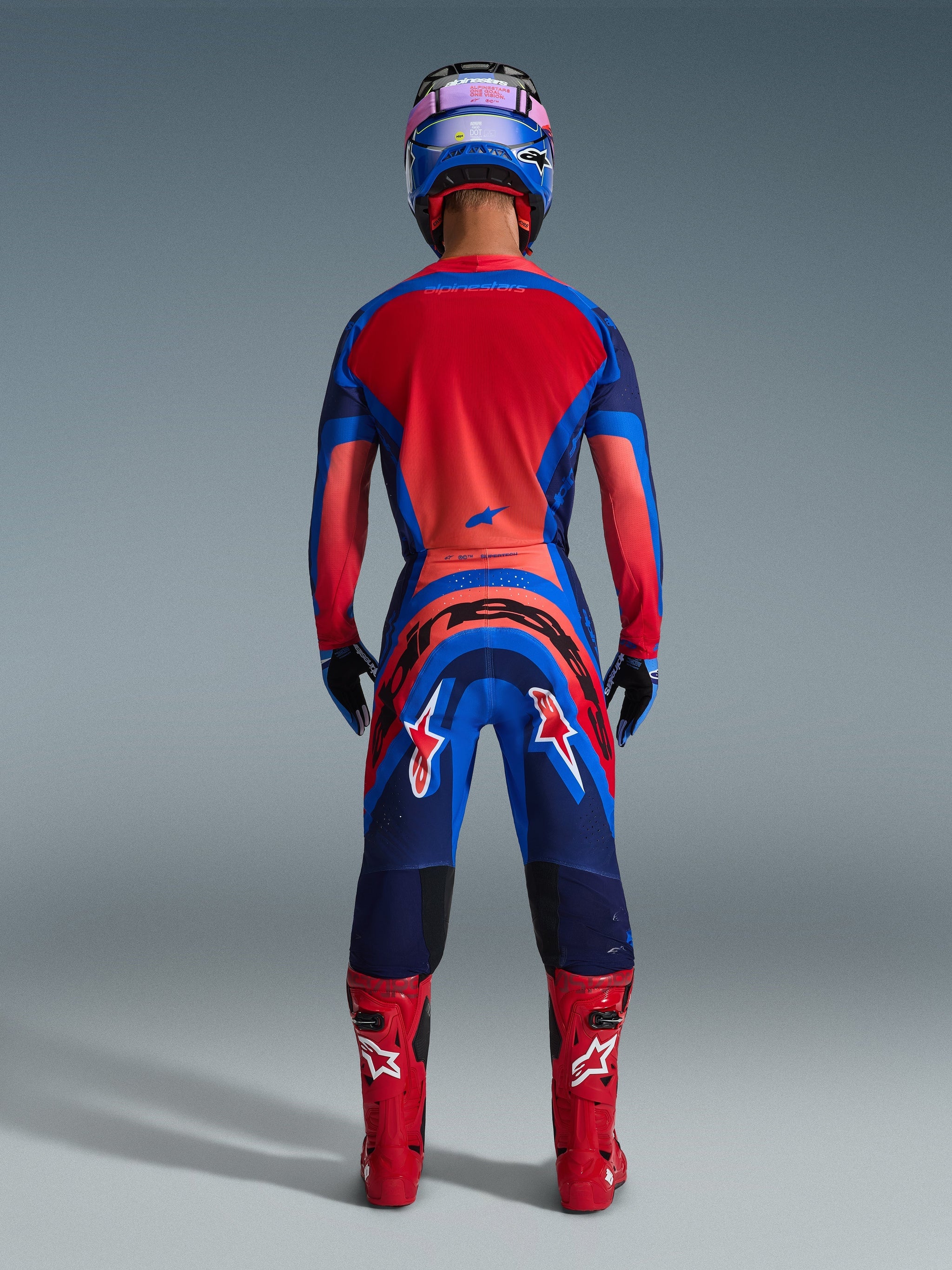 Motocross rider seen from the back wearing the Alpinestars 2026 Supertech Vista Jersey in Dark Navy, Hot Coral, and Cobalt, matching gear set with pants, red racing boots, and a blue helmet, professional off-road racing apparel.