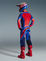 Motocross rider from behind wearing the Alpinestars 2026 Supertech Vista Jersey in Dark Navy, Hot Coral, and Cobalt, paired with matching motocross pants, red boots, and a colorful helmet, coordinated professional racing gear