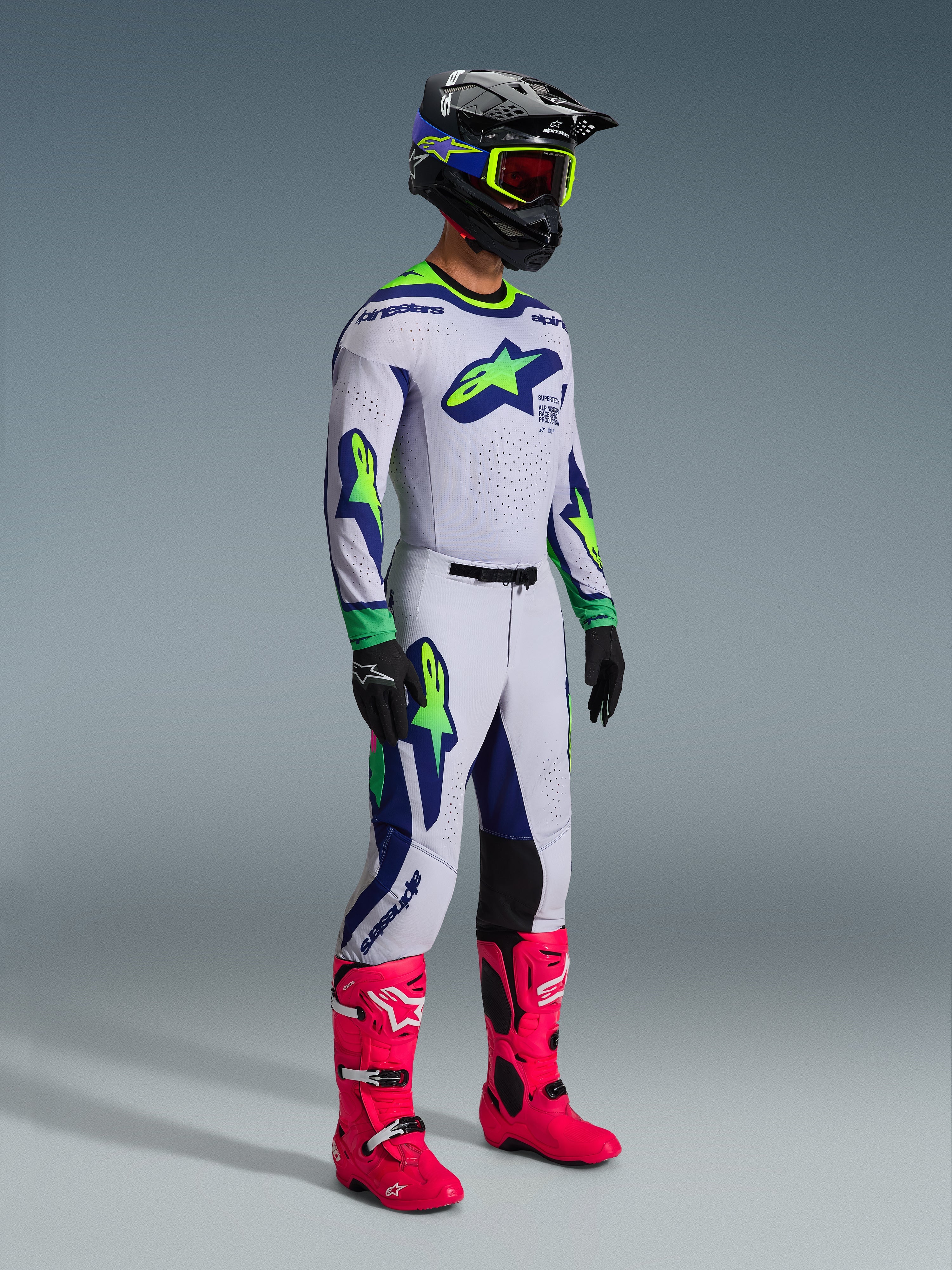 Motocrossförare klädd i Alpinestars 2026 Supertech Vista-tröja i Light Gray Yellow Fluo Blue, matchande racingbyxor, starkt rosa stövlar och en svart hjälm med gula goggles, med laserskuren ventilation och en professionell atletisk passform för terrängkörning