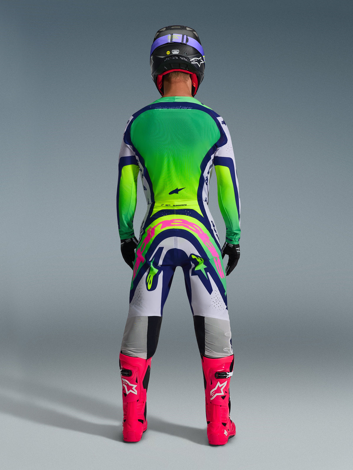 Motocrossförare sedd bakifrån i komplett racingutrustning, 2026 Supertech Vista Jersey med grön och gul färgtoning på ryggen, vita och blå byxor med stor rosa Alpinestars-logotyp, klarrosa stövlar och en svart hjälm med lila accenter, koordinerad professionell outfit för terrängracing.