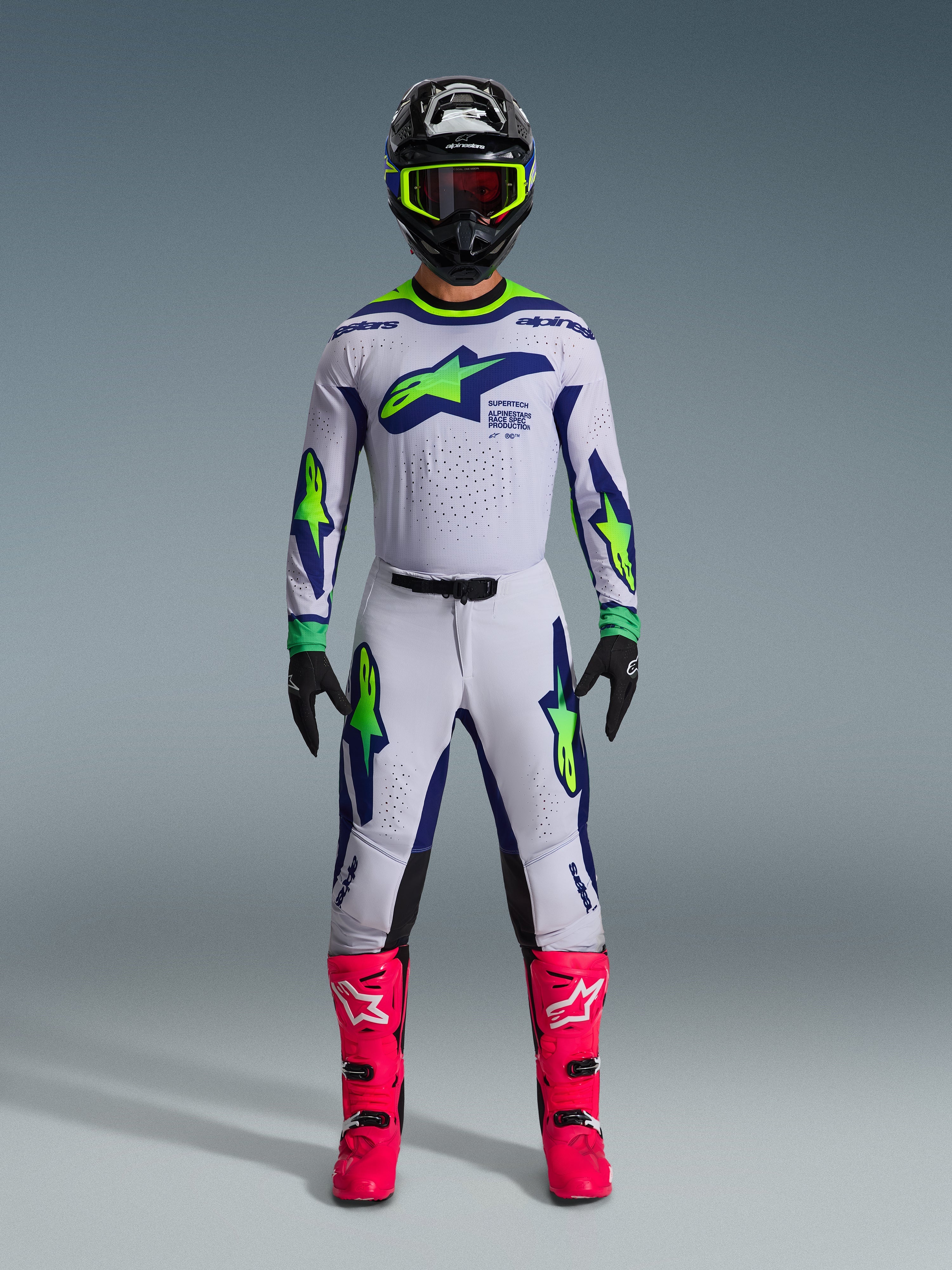 Motocrossförare i 2026 Supertech Vista-tröja i ljusgrått, neongult och blått, matchande byxor, starkt rosa stövlar och en svart hjälm med neondetaljer, off-road racingutrustning på proffsnivå