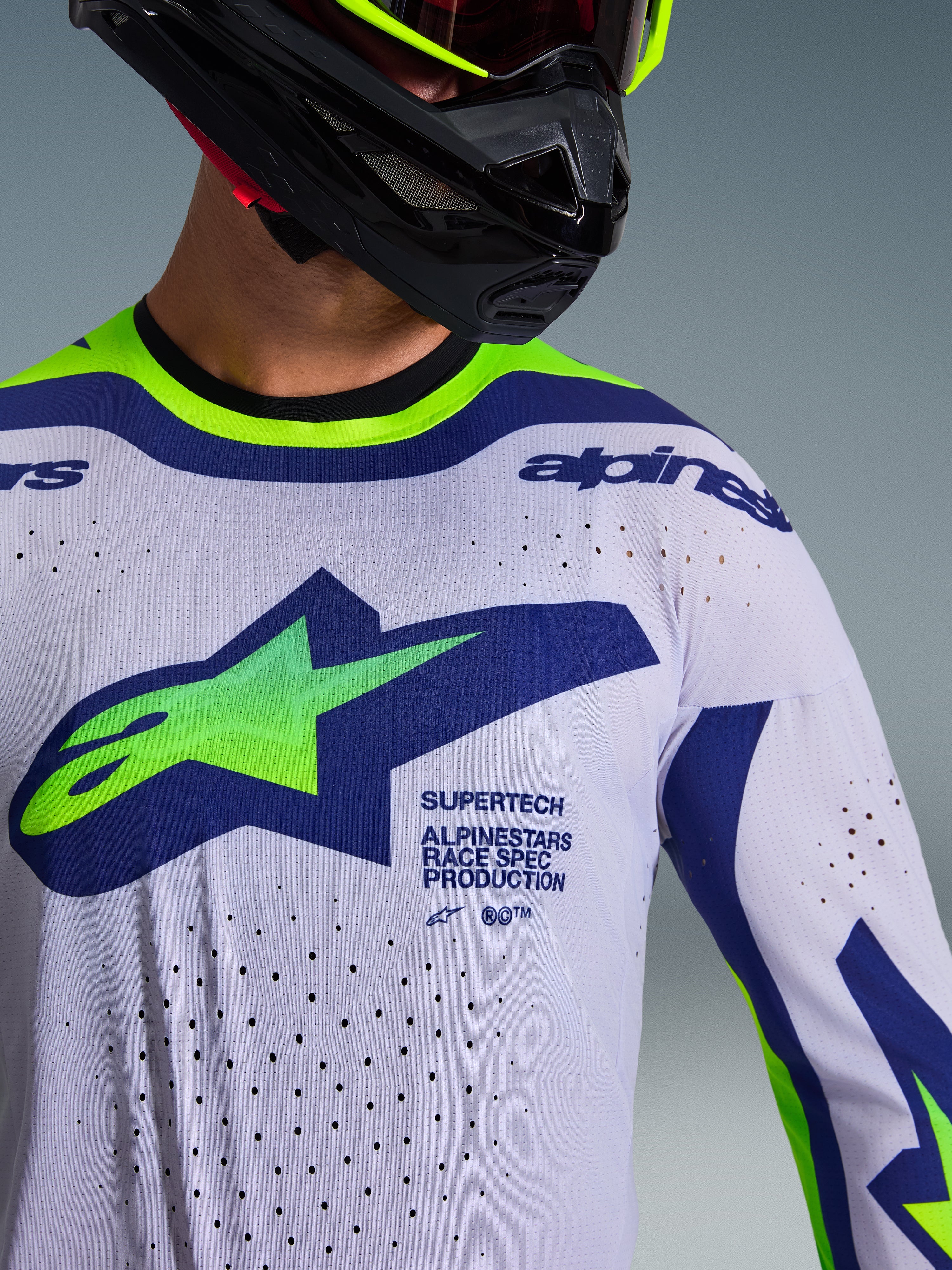 Alpinestars 2026 Supertech Vista-tröja, motocrosströja, ljusgrå med neongula och blå detaljer, bärs av förare med svart hjälm och neongula goggles, har stor grön och blå Astars-logotyp, laserskurna ventilationshål och atletisk race fit-design