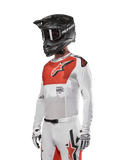 Alpinestars 2024 Supertech Ward-tröja, motocrosströja, vit och Hot Orange, röd bröstpanel med vit stjärnlogotyp, långa ärmar med laserskurna perforeringar, bärs med en svart hjälm och vita handskar, atletisk race-passform