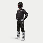 Motocross rider wearing Alpinestars 2024 Fluid Graphite Jersey in Black and Silver, matching black pants with silver star logos, white and black boots, white and black gloves, and a black helmet with red goggles, full off-road racing gear