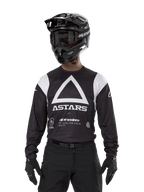 Alpinestars Techdura Jersey, svart, med en stor vit triangelgrafik på bröstet med ASTARS-logotyp och vita axelpaneler, bärs av en förare med svart motocrosshjälm, goggles och matchande svart offroad-utrustning.