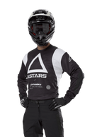 Alpinestars Techdura Jersey, motocrosströja, svart och vit, bärs med matchande hjälm och handskar, pose med händerna på höfterna mot vit bakgrund, med stor triangelformad A-logotyp och ASTARS-branding
