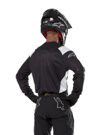 Alpinestars Techdura-tröja, svart med vita axeldetaljer, vy bakifrån på en motocrossförare som bär matchande svarta byxor och hjälm, förstärkta armbågspartier, enduro- och terrängkörningsutrustning