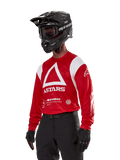 Alpinestars Techdura Jersey, motocrosströja, klarröd och vit, med en framträdande vit triangeldesign och ASTARS-logotyper, bärs med svart hjälm och handskar, slitstark och fukttransporterande utrustning för terrängkörning