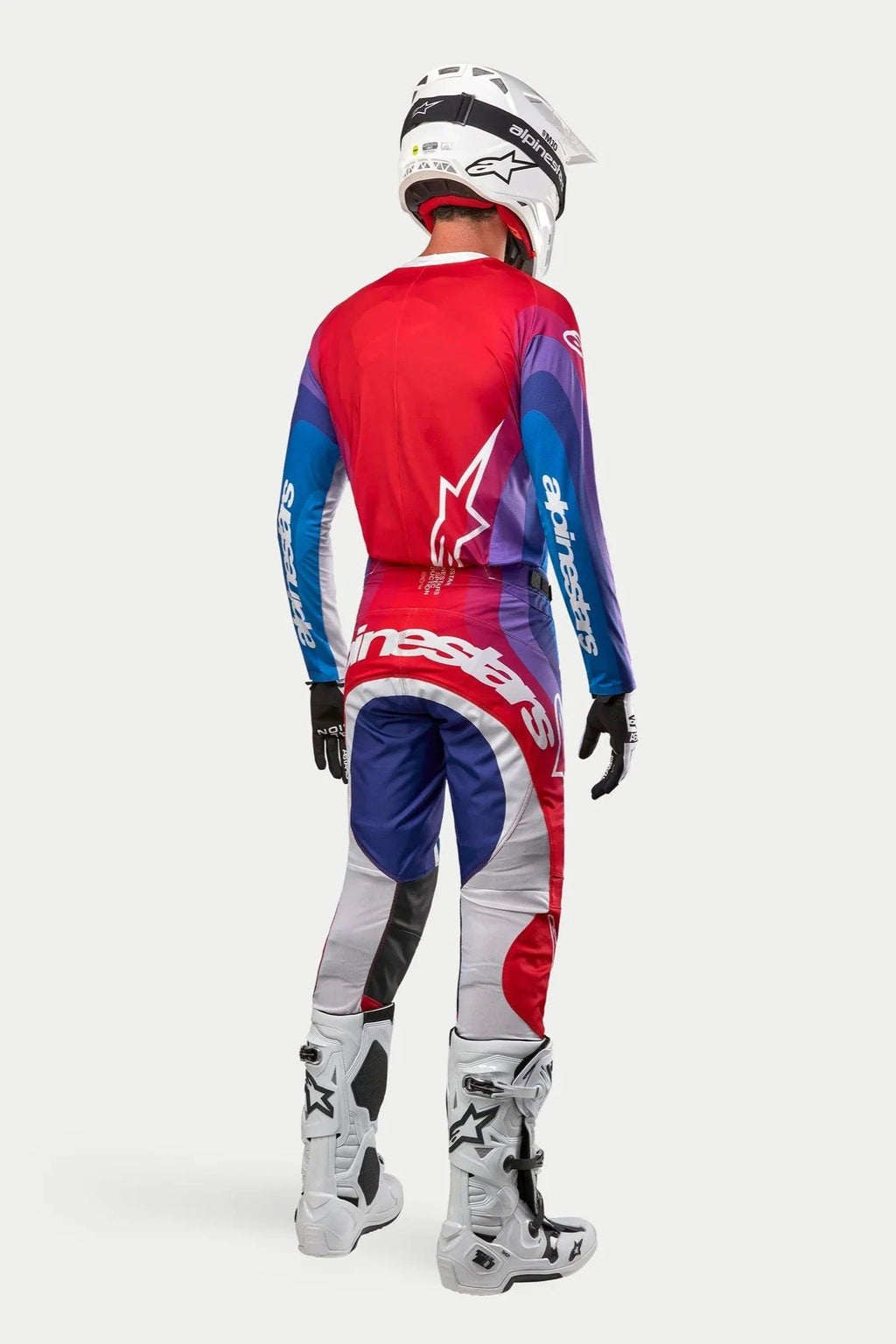Motocrossförare sedd bakifrån i Alpinestars 2024 Techstar Pneuma-tröja och -byxor, färgställningen Blu Mars Red White med röda, blå och lila färgtoningar och vit branding, kombinerat med vita stövlar och en vit hjälm, matchande racingutrustning för terrängkörning.