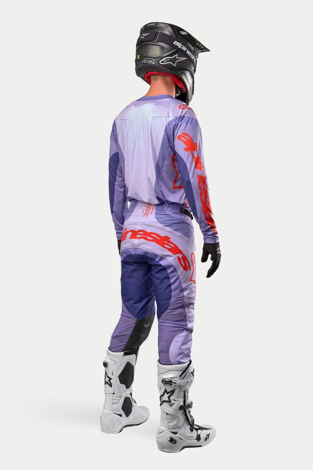 Motocrossförare i komplett racingutrustning sedd bakifrån, lila Alpinestars 2024 Techstar Ocuri-tröja med logotyper i starkt orange, matchande lila byxor med orange detaljer, svart hjälm, vita och svarta stövlar, svarta handskar, professionell utrustning för terrängkörning