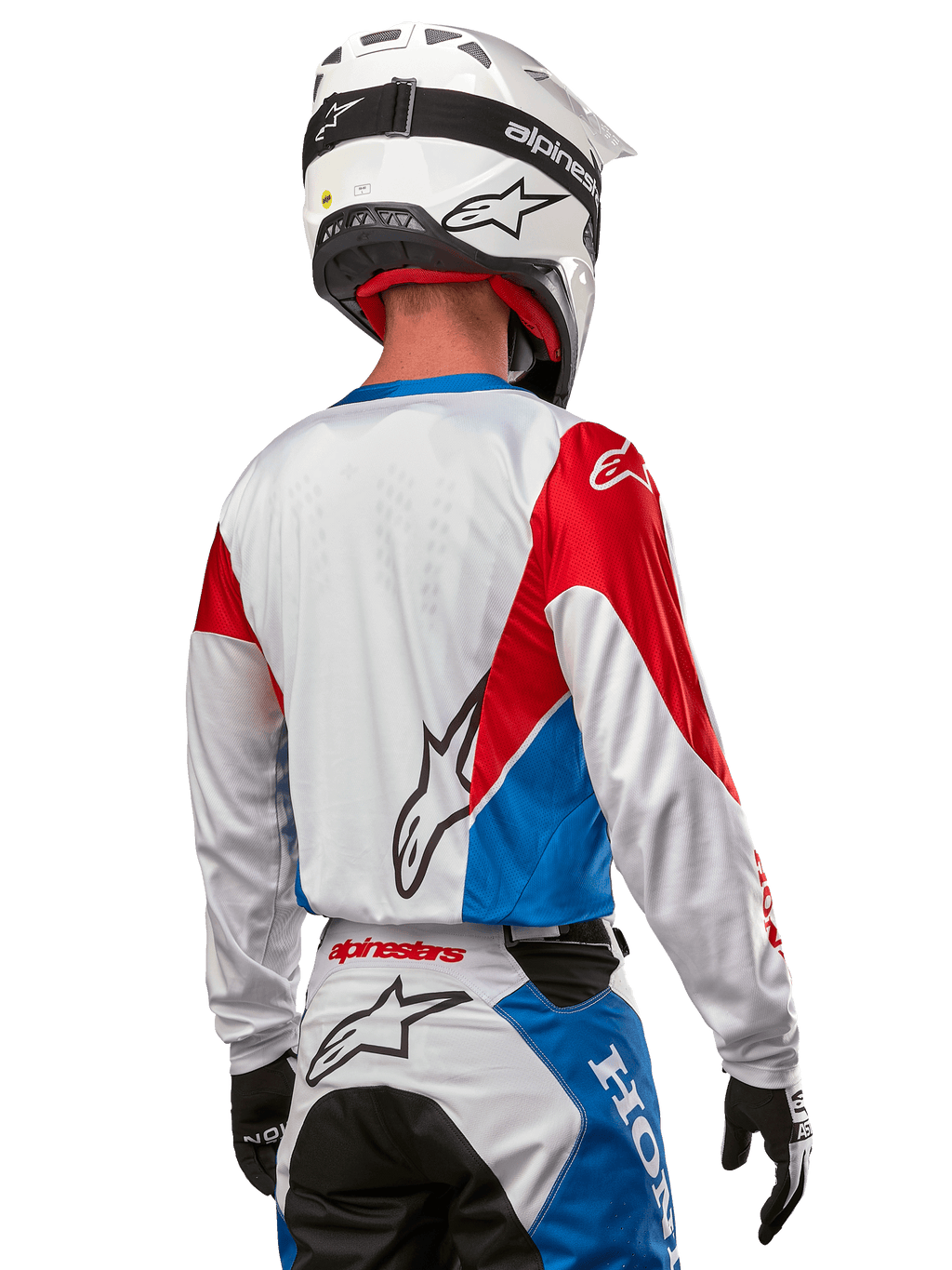 Motocrossförare i Alpinestars Honda Racer Iconic-tröja, sedd bakifrån, vit med klarblå och klarröda paneler, framträdande Alpinestars-logotyp på ryggen, kombinerat med vit motocrosshjälm och matchande racingbyxor.