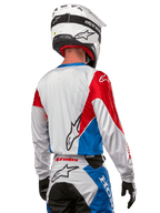 Motocrossförare i Alpinestars Honda Racer Iconic-tröja, sedd bakifrån, vit med klarblå och klarröda paneler, framträdande Alpinestars-logotyp på ryggen, kombinerat med vit motocrosshjälm och matchande racingbyxor.
