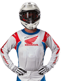 Alpinestars Honda Racer Iconic tröja, vit motocrosströja med röd Honda-logotyp och klarblå och röda detaljer, bärs av en förare med vit hjälm och handskar, prestandautrustning för offroad- och motocrossracing