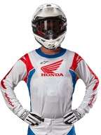 Alpinestars Honda Racer Iconic tröja, vit motocrosströja med röd Honda-logotyp och klarblå och röda detaljer, bärs av en förare med vit hjälm och handskar, prestandautrustning för offroad- och motocrossracing