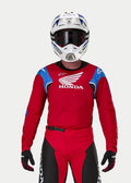 Motocrossförare klädd i Alpinestars Honda Racer Iconic-tröja, klarröd med blå och vita detaljer och Honda-logotyp, matchad med vit och blå hjälm, mörka goggles och matchande röda och svarta motocrossbyxor, prestandautrustning för terrängkörning