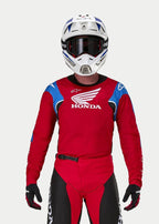 Motocrossförare klädd i Alpinestars Honda Racer Iconic-tröja, klarröd med blå och vita detaljer och Honda-logotyp, matchad med vit och blå hjälm, mörka goggles och matchande röda och svarta motocrossbyxor, prestandautrustning för terrängkörning