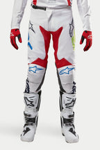Alpinestars 2024 Racer Hana-byxor, motocrossbyxor, vita flerfärgade med röda, blå och limegröna detaljer, bärs med vita och svarta stövlar och röda handskar, med Alpinestars-logotyper på knäna och en hållbar teknisk design för terrängkörning