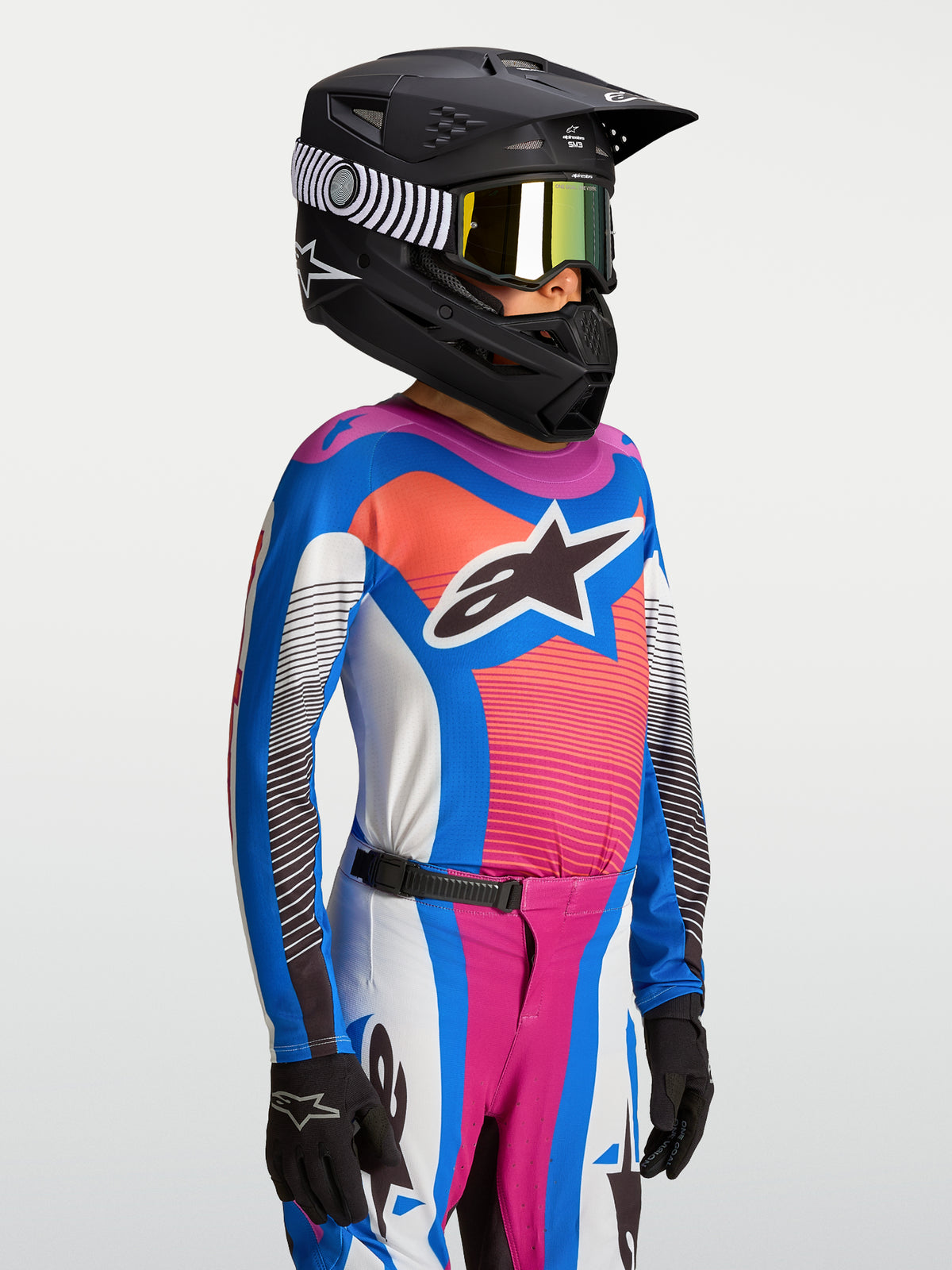 Alpinestars 2027 Supertech Maven ungdomströja, blå och korallrosa andningsbar MX-tröja för terrängkörning, bärs med svart hjälm.
