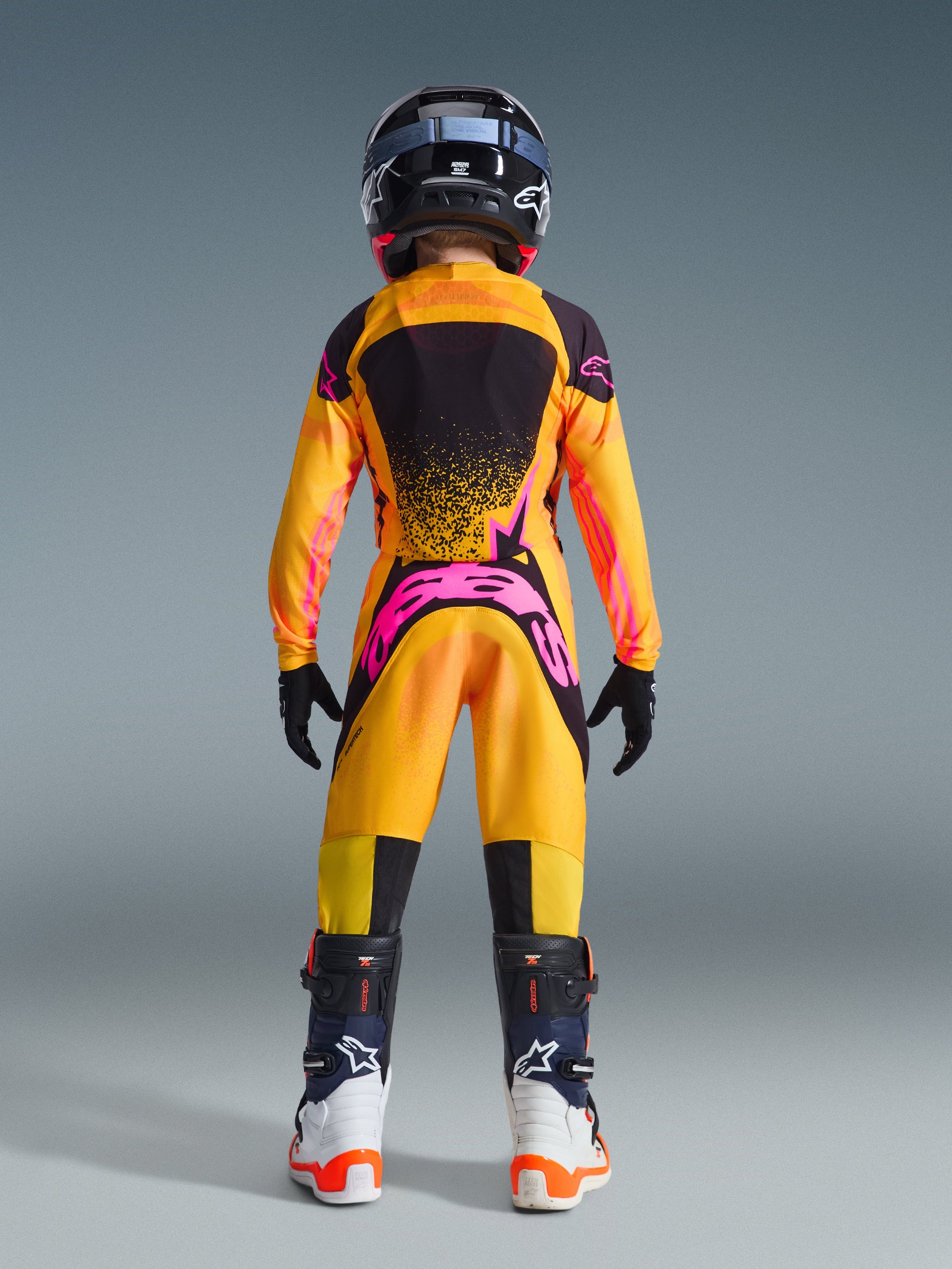 Vy bakifrån av en ungdomsmotocrossförare i Alpinestars 2026 Supertech Nomur-tröja för ungdom, färgkombination i orange, gult och rosa, matchande byxor, svart hjälm och vita och svarta stövlar, högpresterande racingutrustning för unga förare.
