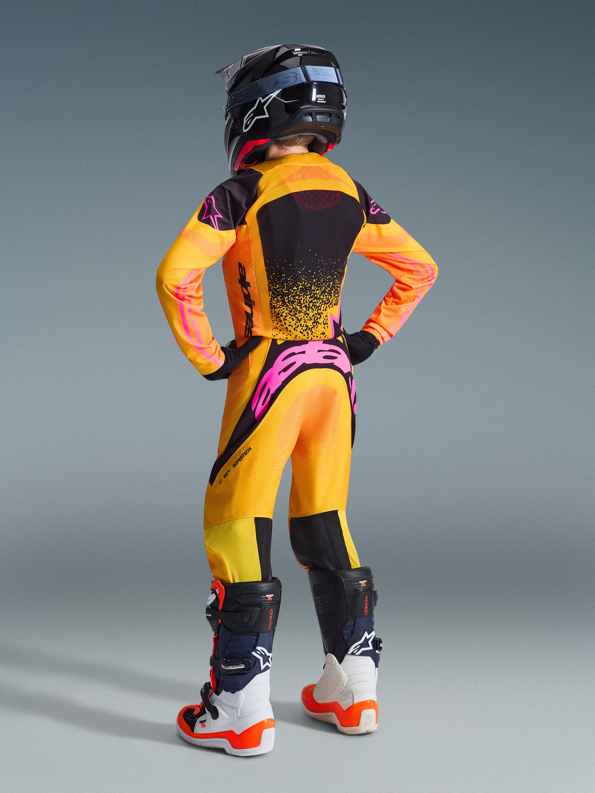 Ungdomsmotocrossförare sedd bakifrån, Alpinestars 2026 Youth Supertech Nomur tröja och matchande byxor i orange, gult och rosa, med livfulla rosa detaljer och svarta meshpaneler, kombinerat med en svart hjälm och stövlar ur Tech-serien, professionell racingutrustning för ungdomar