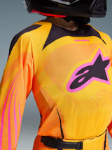 Alpinestars 2026 Youth Supertech Nomur Jersey, motocrosströja i livfull orange, gult och rosa, med en stor svart A-stjärna-logotyp med rosa kant på bröstet, lättviktigt stretch-meshtyg med laserskurna perforeringar, bärs av en förare som en del av en professionell racingutrustning för terrängkörning.
