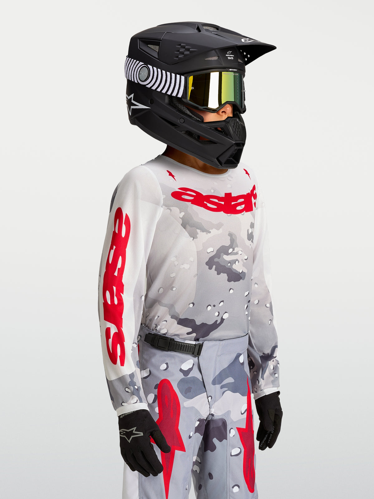 Alpinestars 2027 Ungdom Fluid Lone tröja, grå kamouflagemönstrad och röd motocrosströja på modell med hjälm.