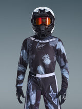Alpinestars 2026 Youth Fluid Lahnd-tröja, motocrosströja, Iron Camo, buren av en ungdomsförare med matchande byxor, svart och vit hjälm med orange goggles och svarta handskar, grå och svart stänkmönstrad design med vit logotyp, komplett offroad-racingutrustning för unga förare.