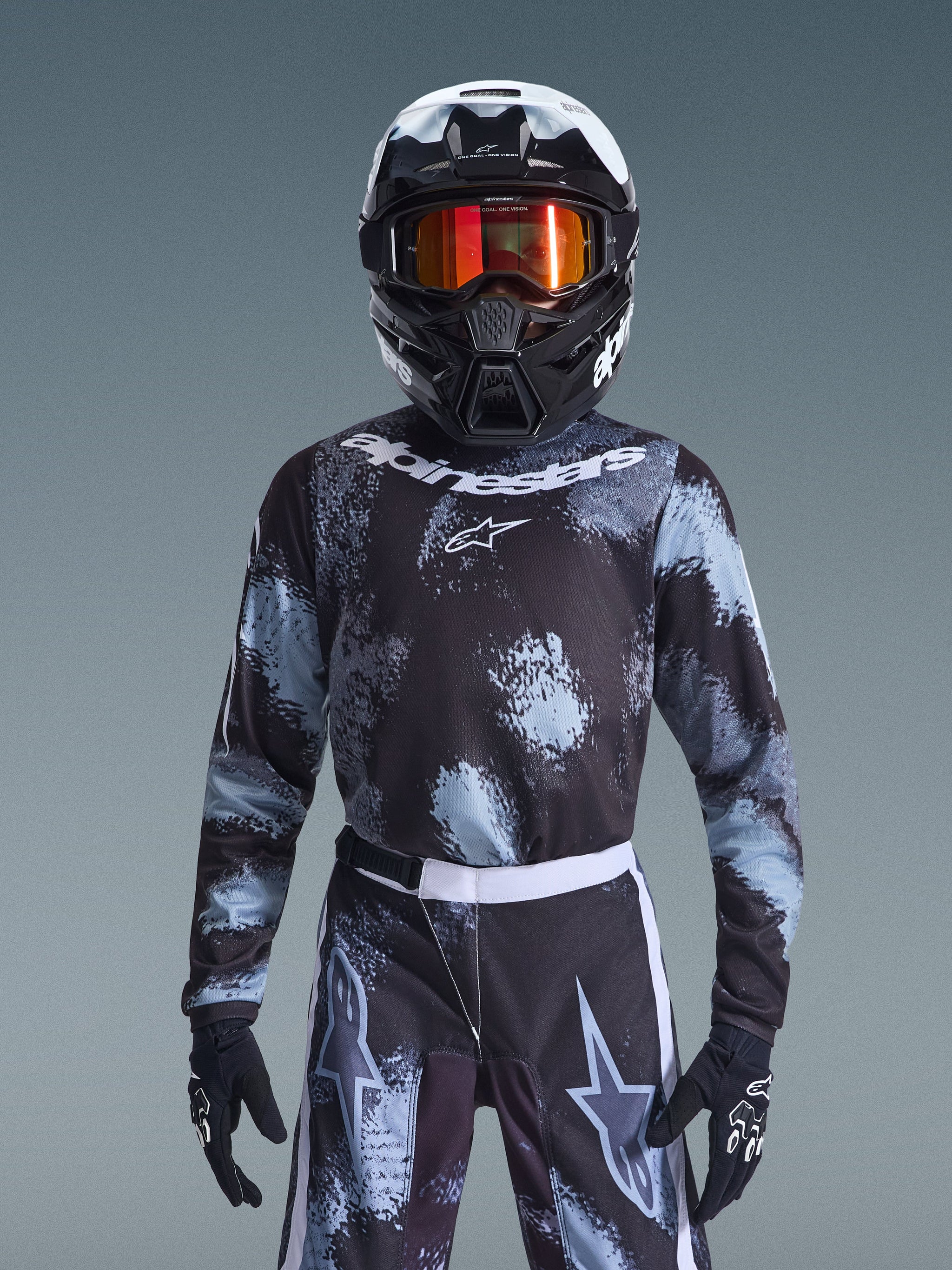 Alpinestars 2026 Youth Fluid Lahnd-tröja, motocrosströja, Iron Camo, buren av en ungdomsförare med matchande byxor, svart och vit hjälm med orange goggles och svarta handskar, grå och svart stänkmönstrad design med vit logotyp, komplett offroad-racingutrustning för unga förare.