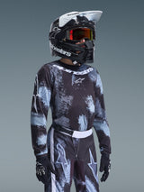 Alpinestars 2026 Ungdom Fluid Lahnd-tröja, motocrosströja, Iron Camo-mönster i svart och ljusblått, buren av en ung förare med matchande byxor och svart hjälm, koordinerad racingutrustning för terrängkörning.