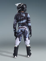 Vy bakifrån av en ungdomsförare iklädd Alpinestars 2026 Ungdom Fluid Lahnd-tröja och matchande byxor i Iron Camo, med ett svart och grått kamouflagemönster med vita detaljer, matchat med en svart och vit hjälm och svarta motocross-stövlar, komplett offroad-racingutrustning för ungdomar.