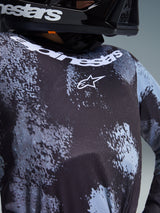Närbild på Alpinestars 2026 Youth Fluid Lahnd-tröja i Iron Camo, med ett svart och grått kamouflagemönster, vit Alpinestars-logotyp på bröstet och märkesnamn längs halsringningen, buren av en förare i en svart hjälm.