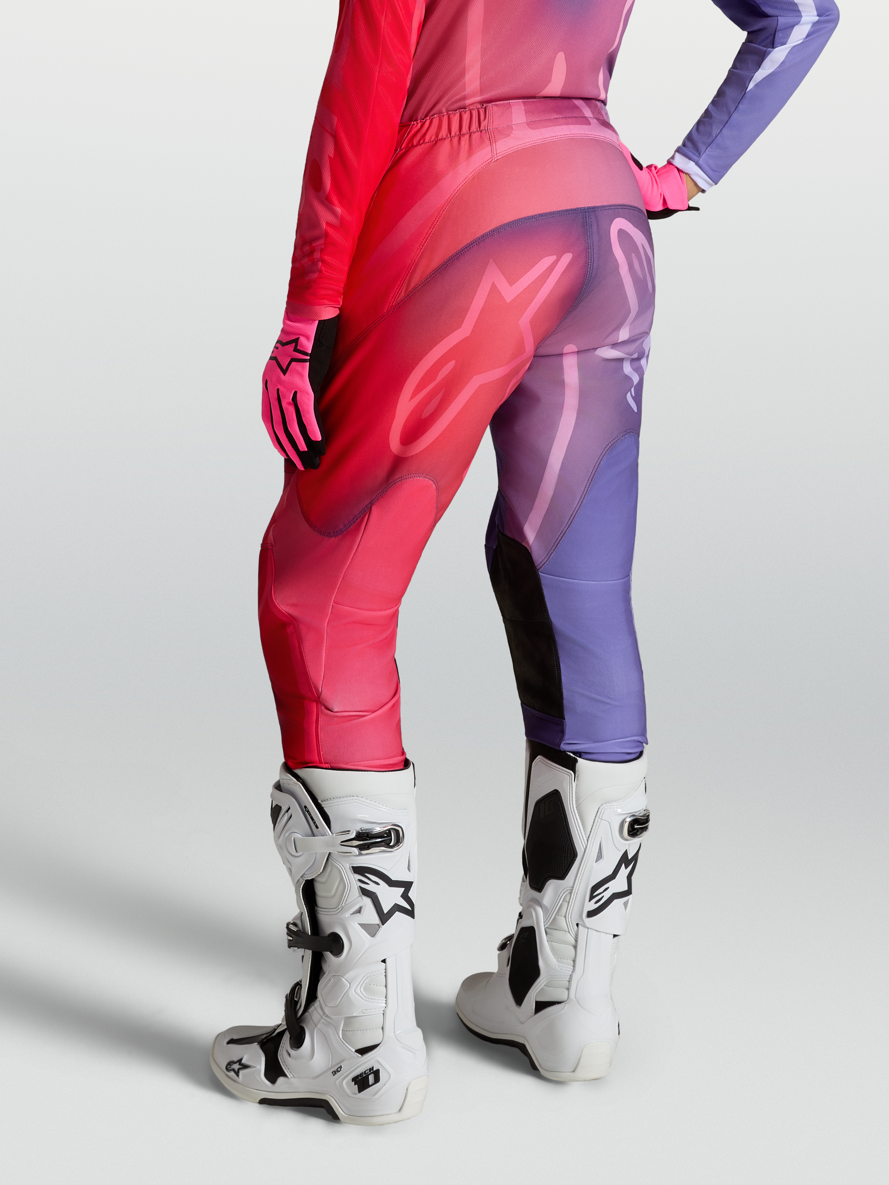Alpinestars Stella Fluid Kepler byxor, motocrossbyxor för dam, lila och röda, vy bakifrån med förböjda ben och stretchpaneler, på modell med vita Tech 10 stövlar.