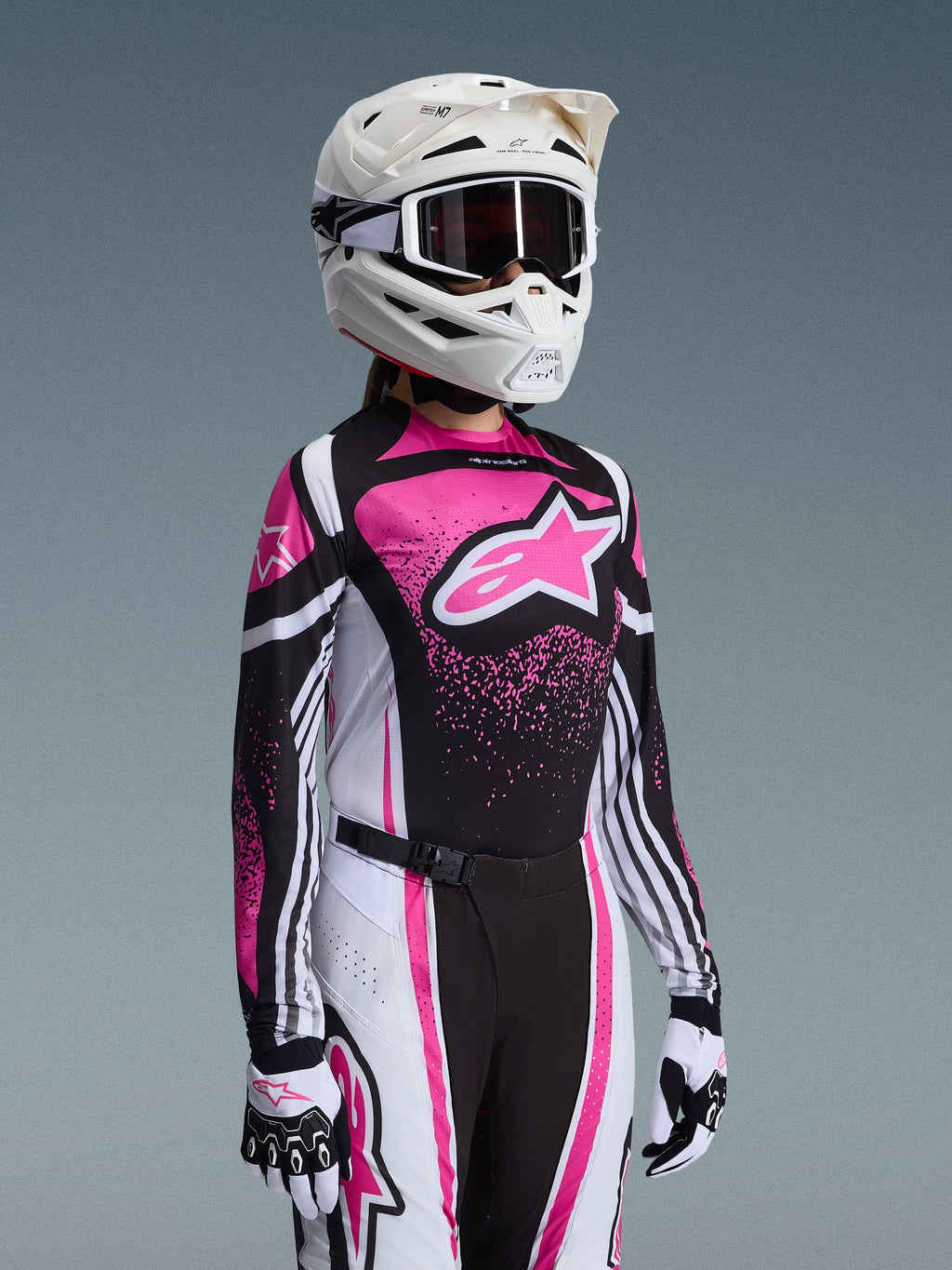 Alpinestars 2026 Stella Techstar Nomur-utrustningsset, kvinnlig motocrossförare i rosa, svart och vitt, tröja med en stor rosa stjärnlogotyp, matchande byxor med magnetiskt spänne, vit hjälm med goggles, vita och svarta handskar.