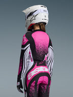 Kvinnlig motocrossförare sedd bakifrån bärande 2026 Stella Techstar Nomur-utrustningsset, rosa, svart och vit tröja med splattergrafik, matchande byxor med stor vit logotyp, vit motocrosshjälm med svarta detaljer, koordinerad professionell racingutrustning