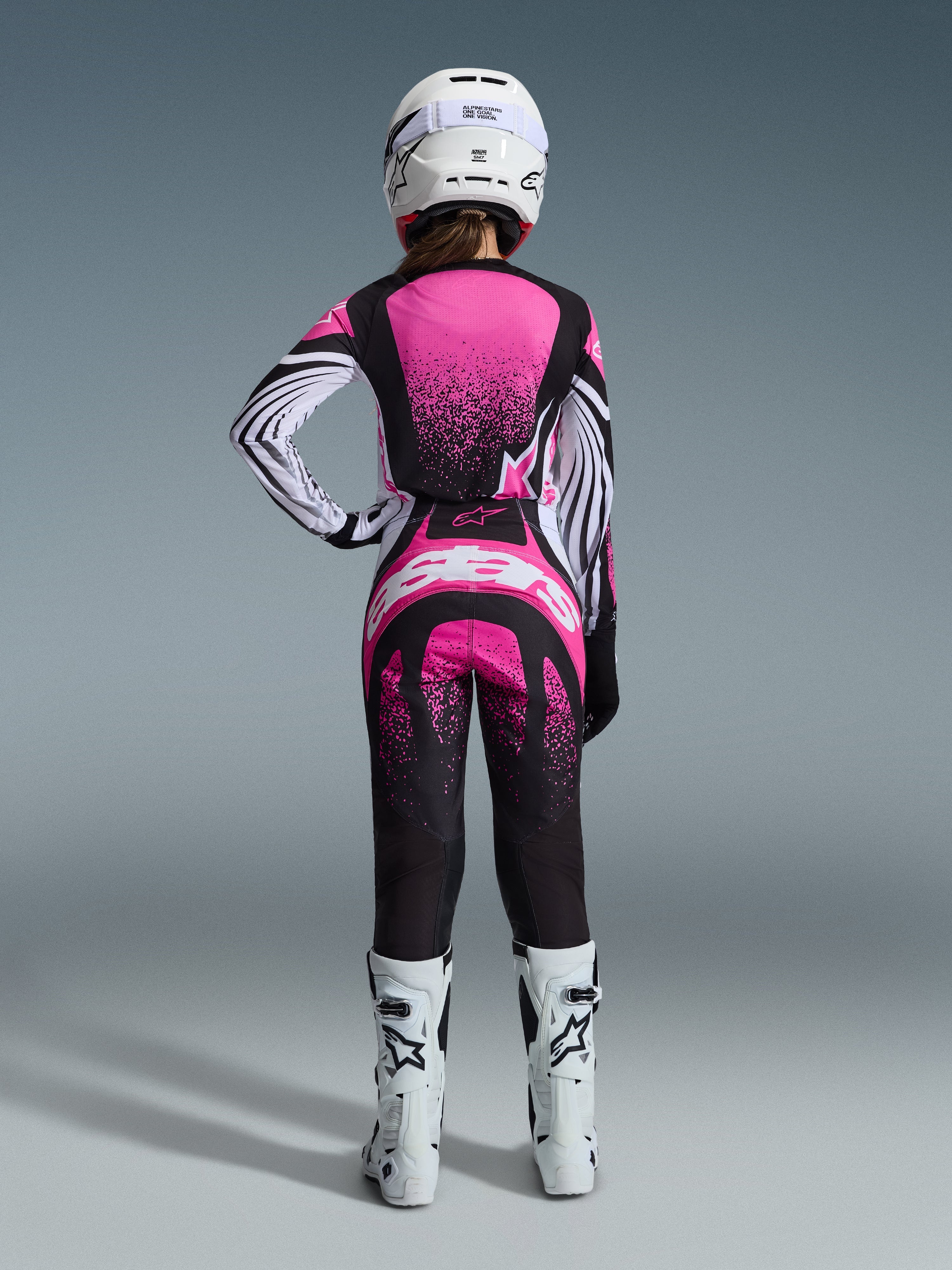 Alpinestars 2026 Stella Techstar Nomur-utrustningsset, kvinnlig motocrossförare sedd bakifrån, rosa och svart tröja med vitrandiga ärmar och spräckligt mönster, matchande byxor med vit Alpinestars-logotyp, vit hjälm och vita offroad-stövlar