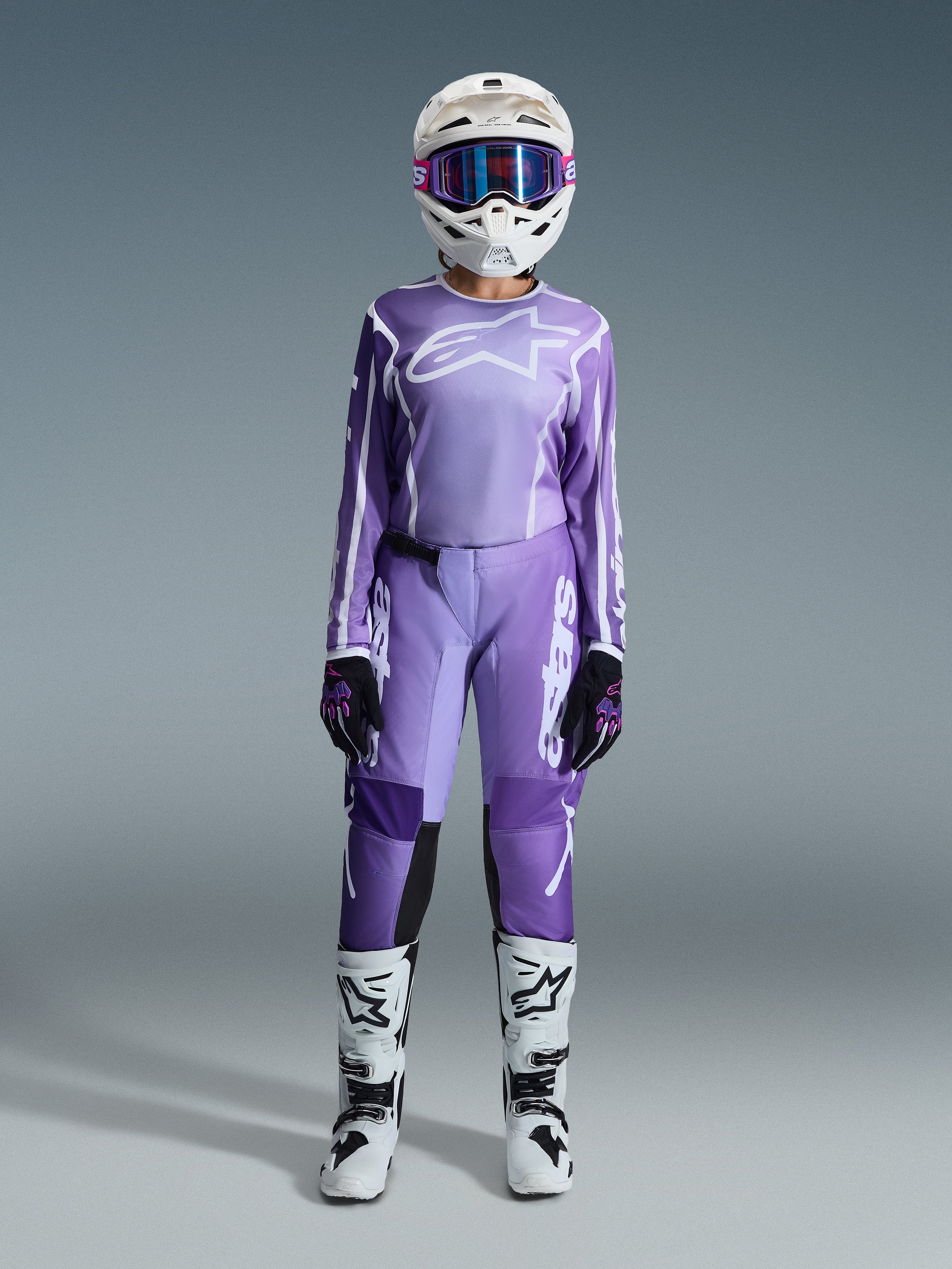 Motocross rider wearing the Alpinestars 2026 Stella Fluid Apex Gear Set, purple jersey with white logo, matching purple pants with white branding, white helmet with pink goggles, white and black boots, coordinated women's motocross outfit for off-road racing